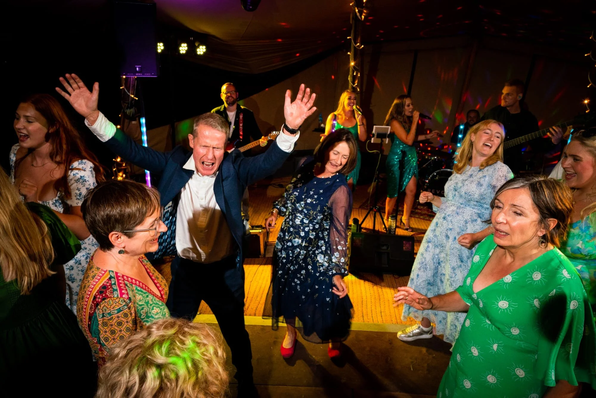 People dancing and enjoying music at a lively party or celebration, with a band performing in the background and colorful lights overhead.