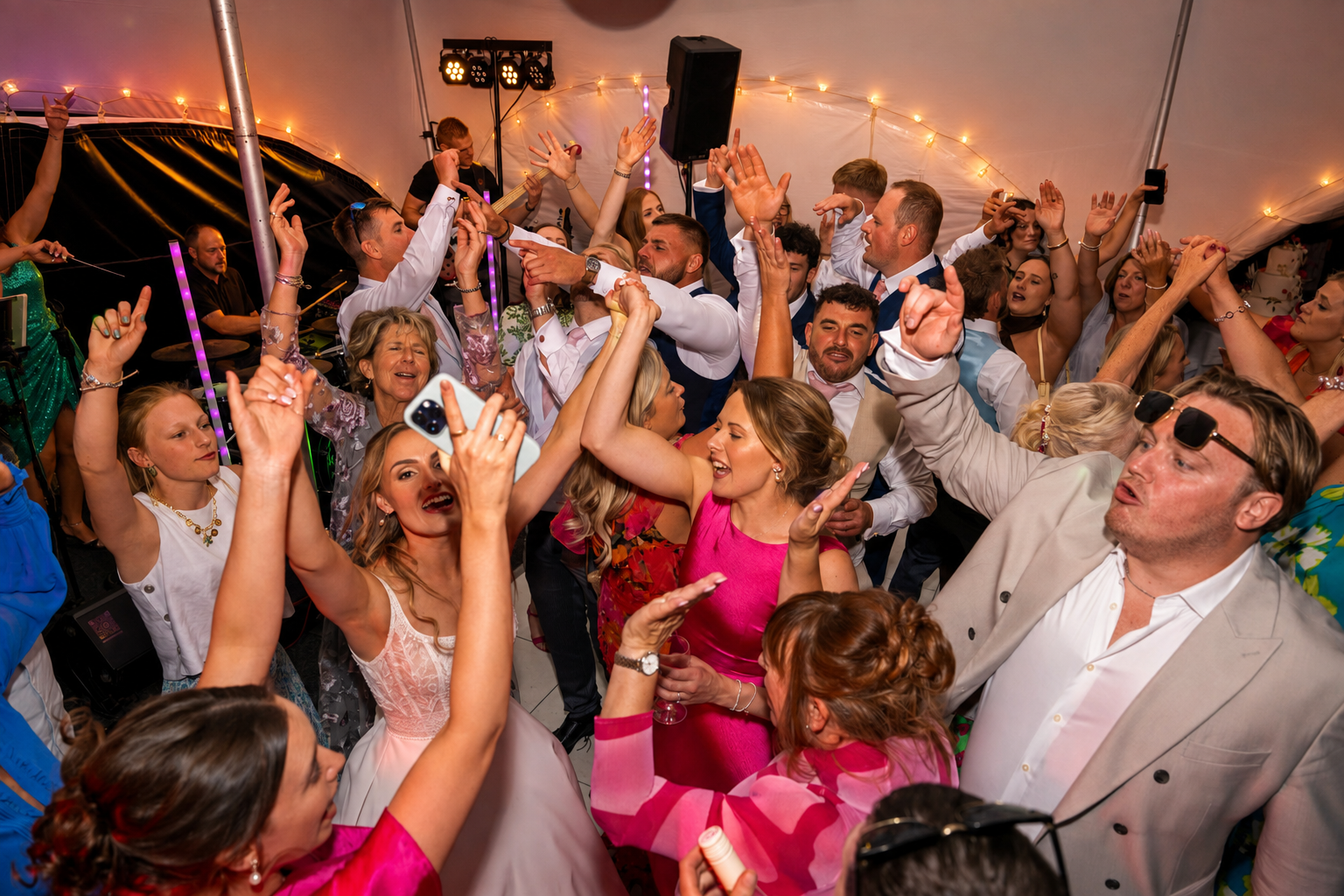 People dancing and celebrating at a wedding or party under string lights, with a DJ playing music in the background.