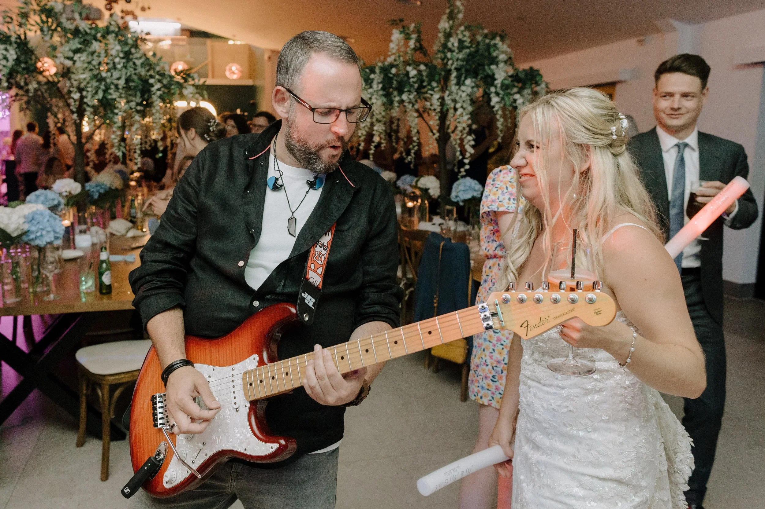 A man wearing glasses and a black shirt is playing an electric guitar, and a woman in a white lace dress is holding a guitar and smiling. They are at a celebration with floral decorations on the tables and trees in the background, with other guests in attendance.