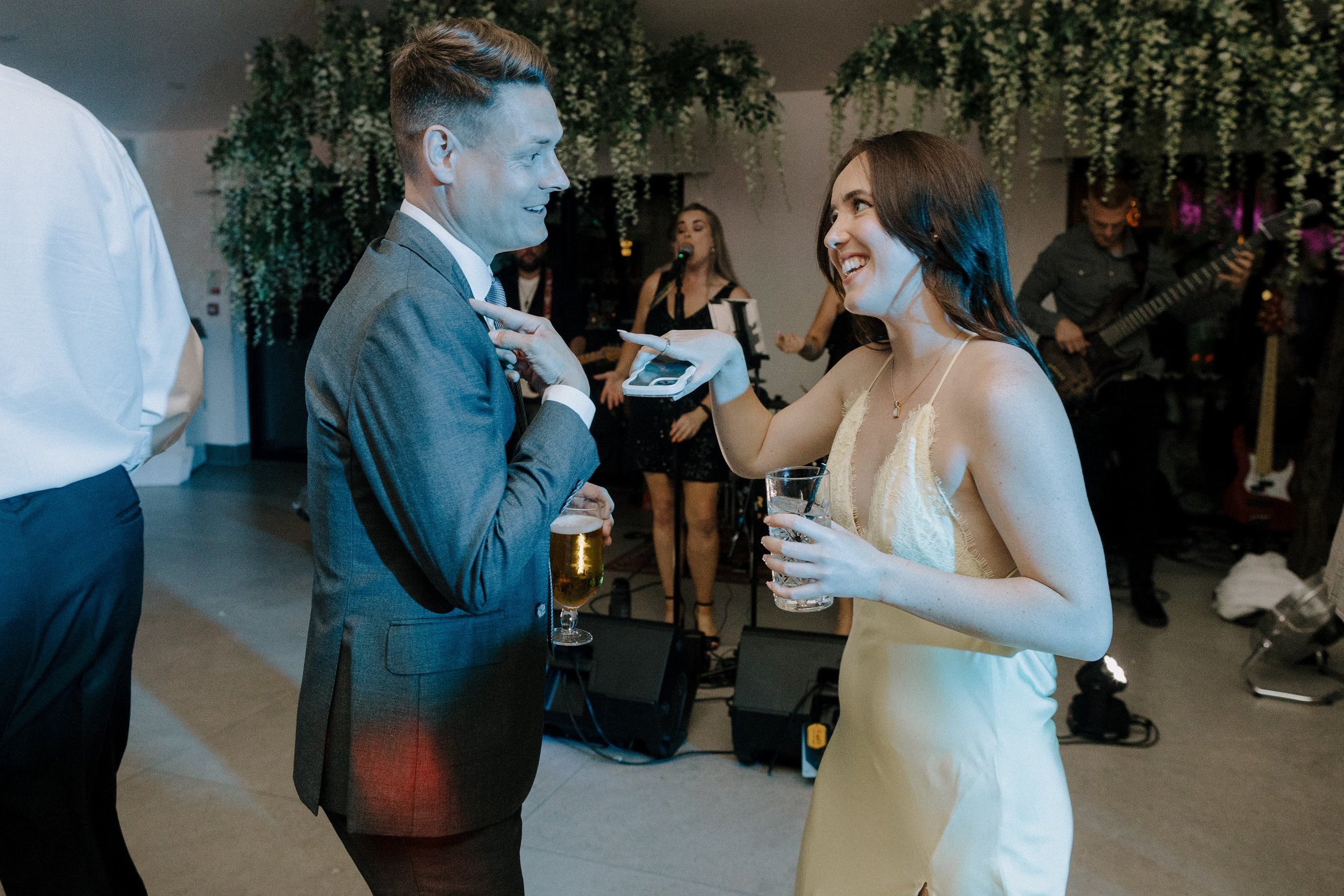 A man and woman dancing at a party. The woman is holding a glass of water, and the man is holding a beer. A band is playing in the background with a woman singing and a man playing the guitar.