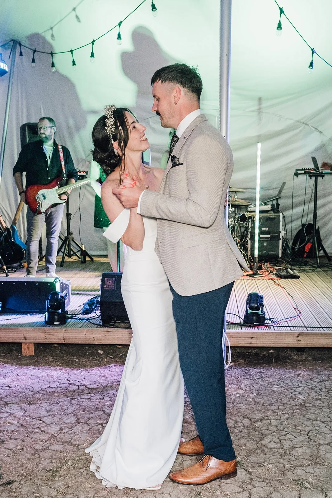 A bride and groom sharing a dance at their wedding reception, with live band playing in the background under string lights.