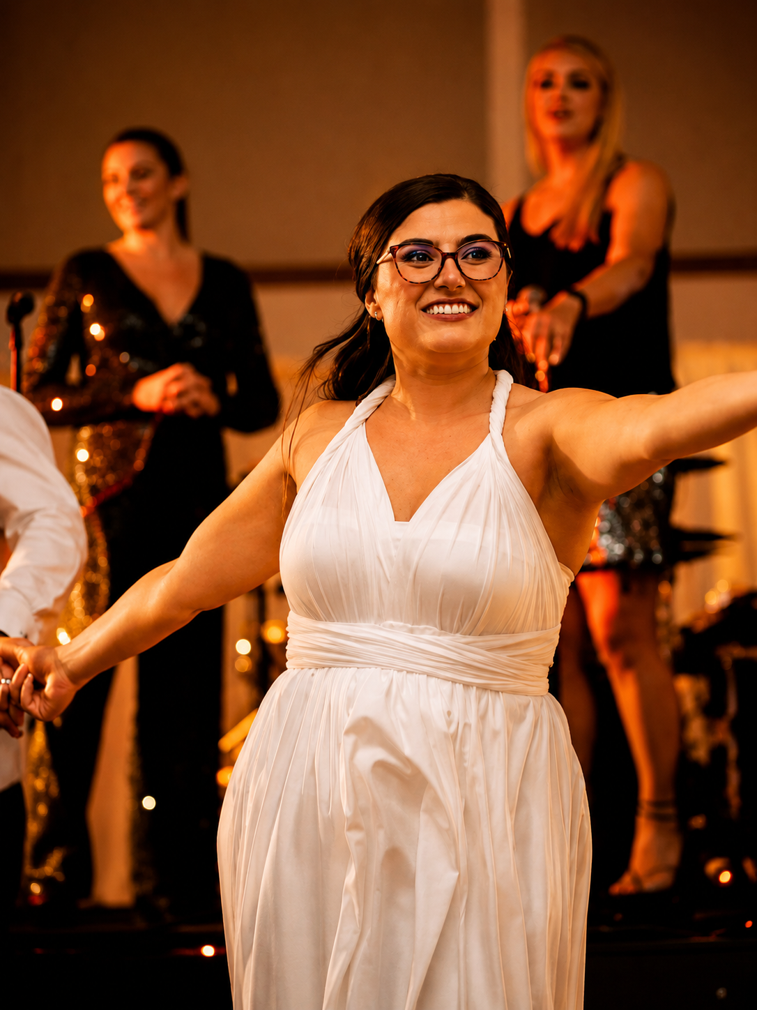 A woman in a white dress with glasses smiling and extending her arm, during a celebration or event, with three women in black dresses in the background.