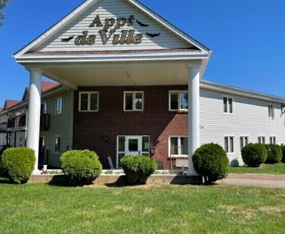 Exterior of a residential building with a sign that reads "Appt de Ville" on the gable end, surrounded by green bushes and a grassy lawn under a clear blue sky.