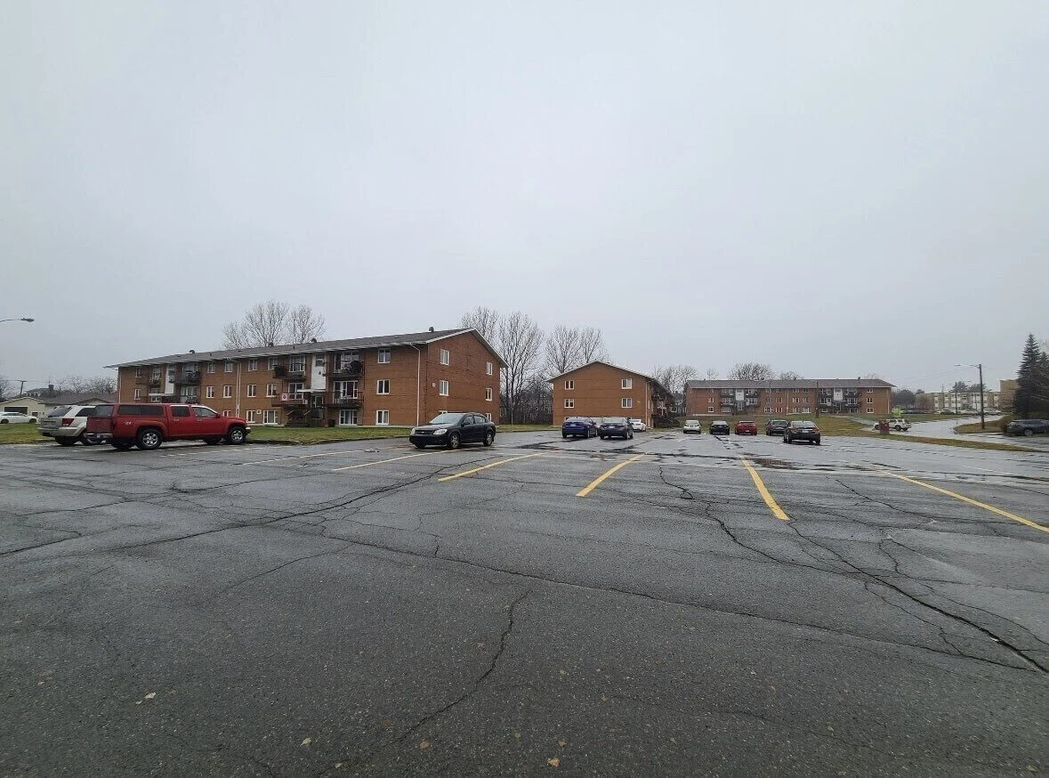 A parking lot with several parked cars and apartment buildings in the background on a cloudy, rainy day.