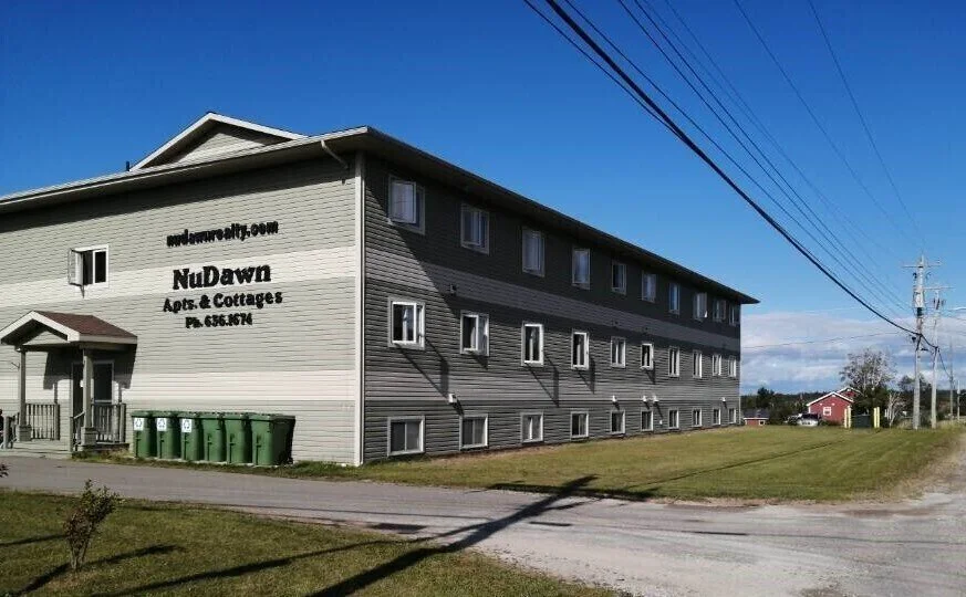 Three-story gray apartment building with a small front porch and green trash bins outside, in a rural area with power lines and a grassy field, under a clear blue sky.
