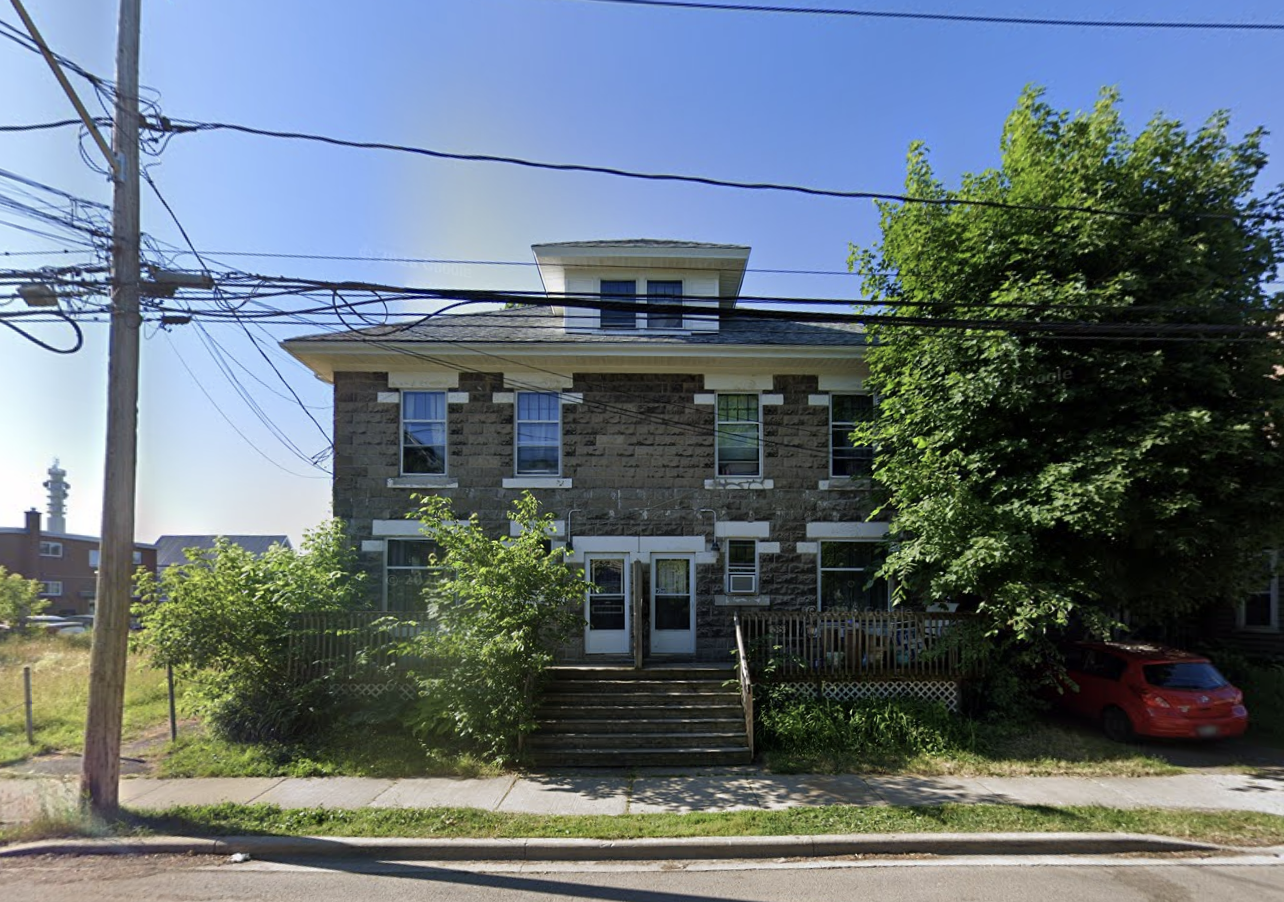 A two-story stone house with a small upper dormer window, a front staircase, and surrounded by green trees and bushes. There are power lines overhead and a red car parked to the right.