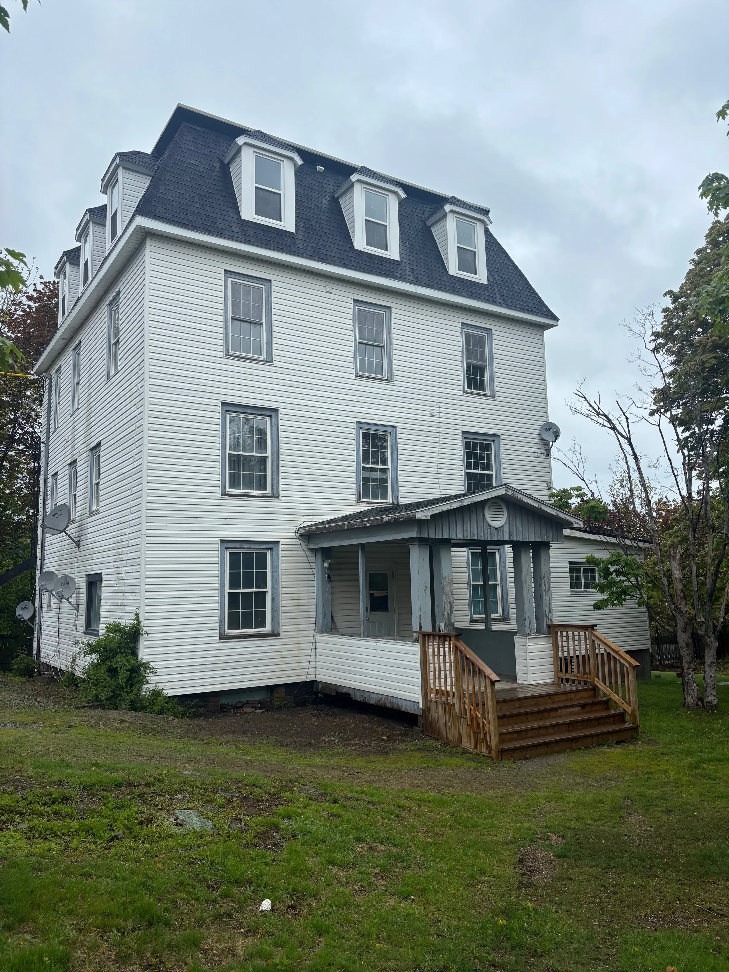 Side view of a white, multi-story house with a large, steep, blue roof and multiple windows. There is a small wooden deck with steps leading up to a porch.