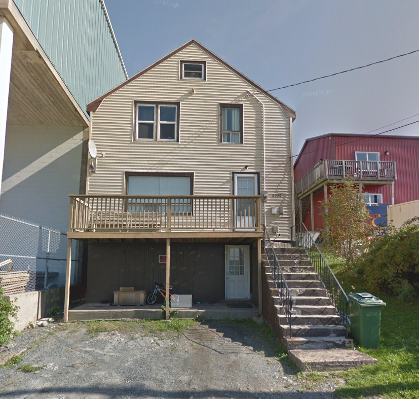 A three-story house with beige vinyl siding, a front porch, and a staircase on the right. The house has multiple windows, a small balcony, and a garage at the bottom. There is a bicycle and other items under the porch.