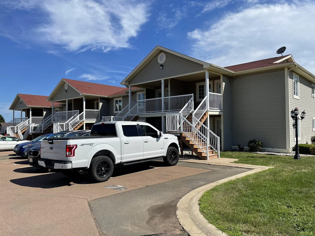 Multi-unit residential building with balconies, parked white truck, and several cars on the driveway under a blue sky with clouds.