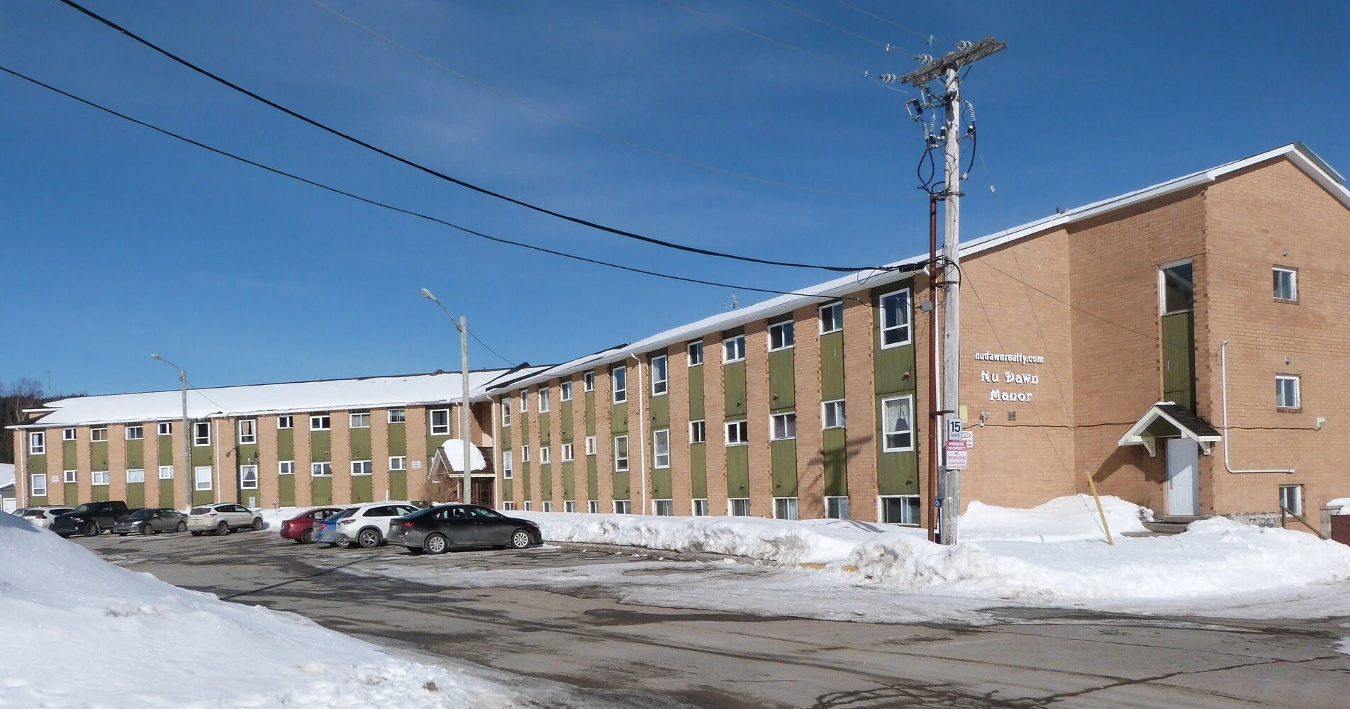 A multi-story brick apartment building with green accents and a snowy parking lot in front. Several cars are parked, and power lines run above. Clear blue sky overhead.