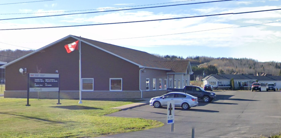 A church building with a Canadian flag, a parking lot with several cars, a sign on a post with a cross, and a rural landscape in the background.