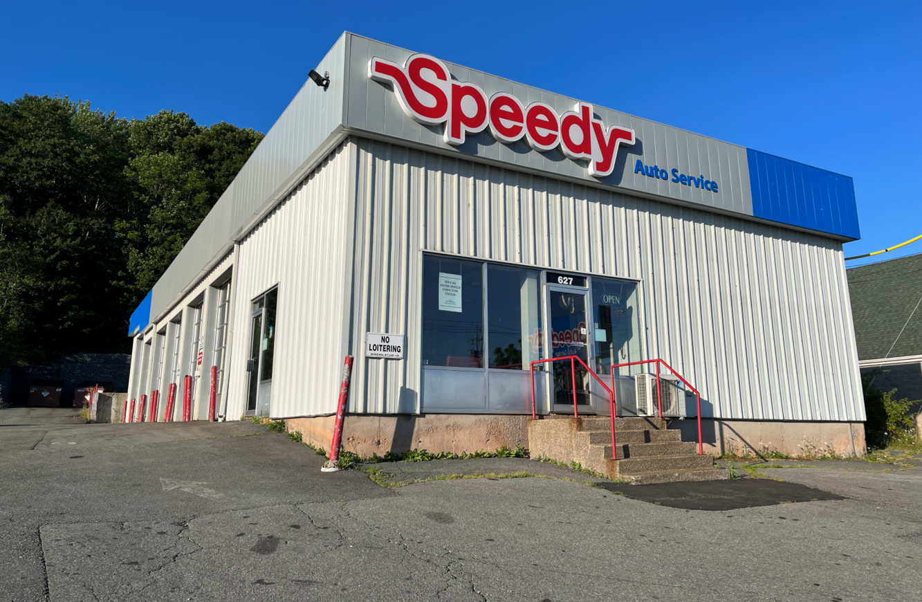 An auto service shop named Speedy with a corrugated metal exterior and a blue awning, located at 627, with a glass door and stairs, no loitering sign, and surrounded by a parking lot and trees.
