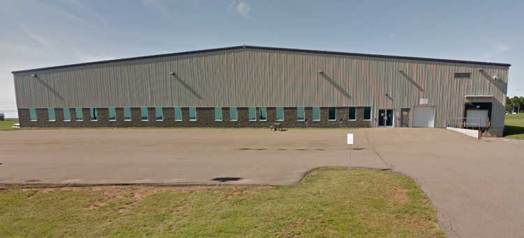 Large industrial warehouse building with a long row of windows, located in a spacious paved area with some grass in the foreground.