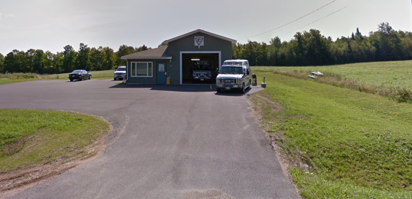A small blue garage with a few parked cars in front and a van nearby, situated on a paved driveway surrounded by green fields and trees.