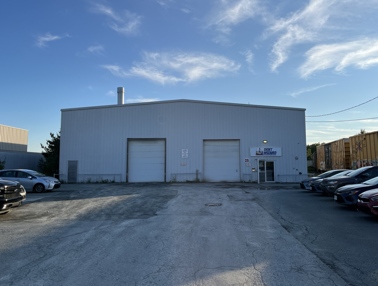 Exterior view of a warehouse building with a sign that reads 'Dent Wizard' and several parked cars in the lot.