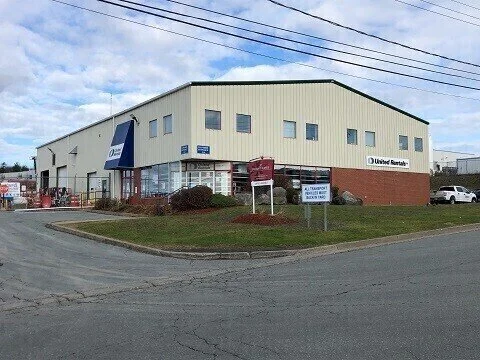 A two-story commercial building with a yellow exterior, blue awning, and signs indicating it is a United Rentals location, situated on a corner lot with a parking area.