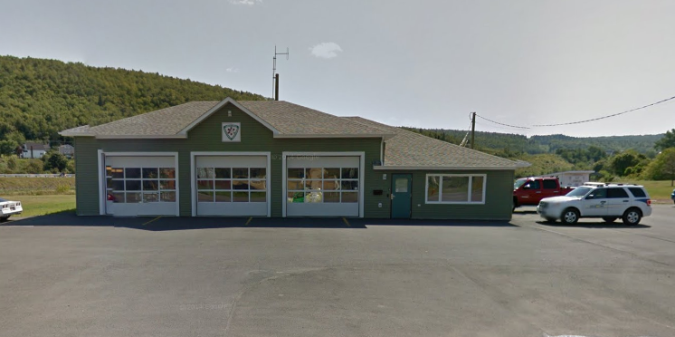 A small, green building with three large garage doors and a parking lot in front. There is a sign with a crest above the garage doors, and several cars parked nearby.