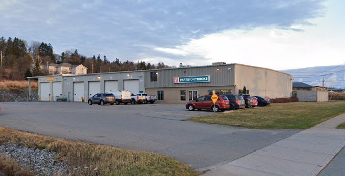 A commercial building with a sign that reads 'Parts for Trucks'. Several cars are parked in front, with a sidewalk and grass area nearby. The sky is partly cloudy.