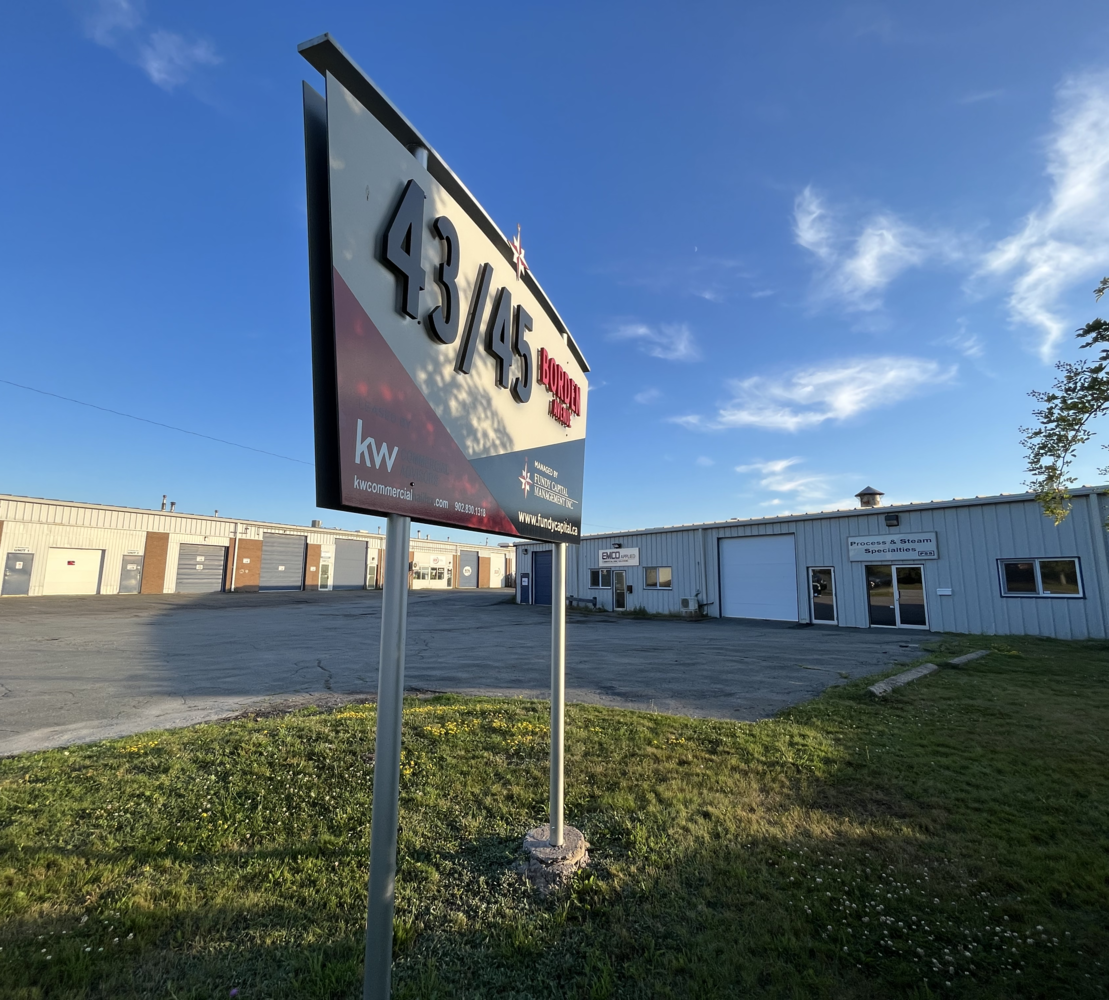 A parking lot with a large sign displaying 43/45 Brooked Avenue in front of a warehouse building under a blue sky.