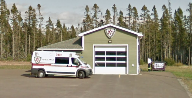 An ambulance parked outside a green fire station building with a large garage door, surrounded by trees and an overcast sky.