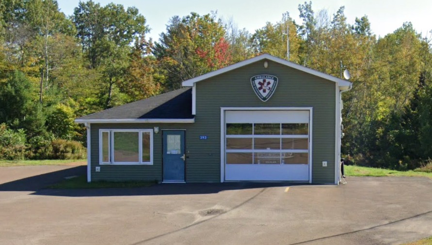 A building with a sign for emergency medical services, featuring a garage door and a side door, surrounded by trees.