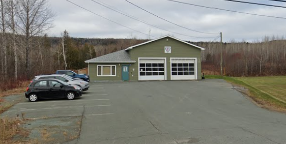 A small green fire station with two garage doors and parking spaces in front, located in a rural area with open fields and trees.