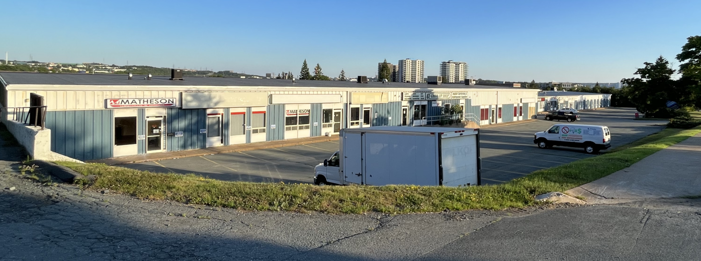 Industrial building with parking lot and a few trucks, including one large box truck. Some high-rise buildings in the background.