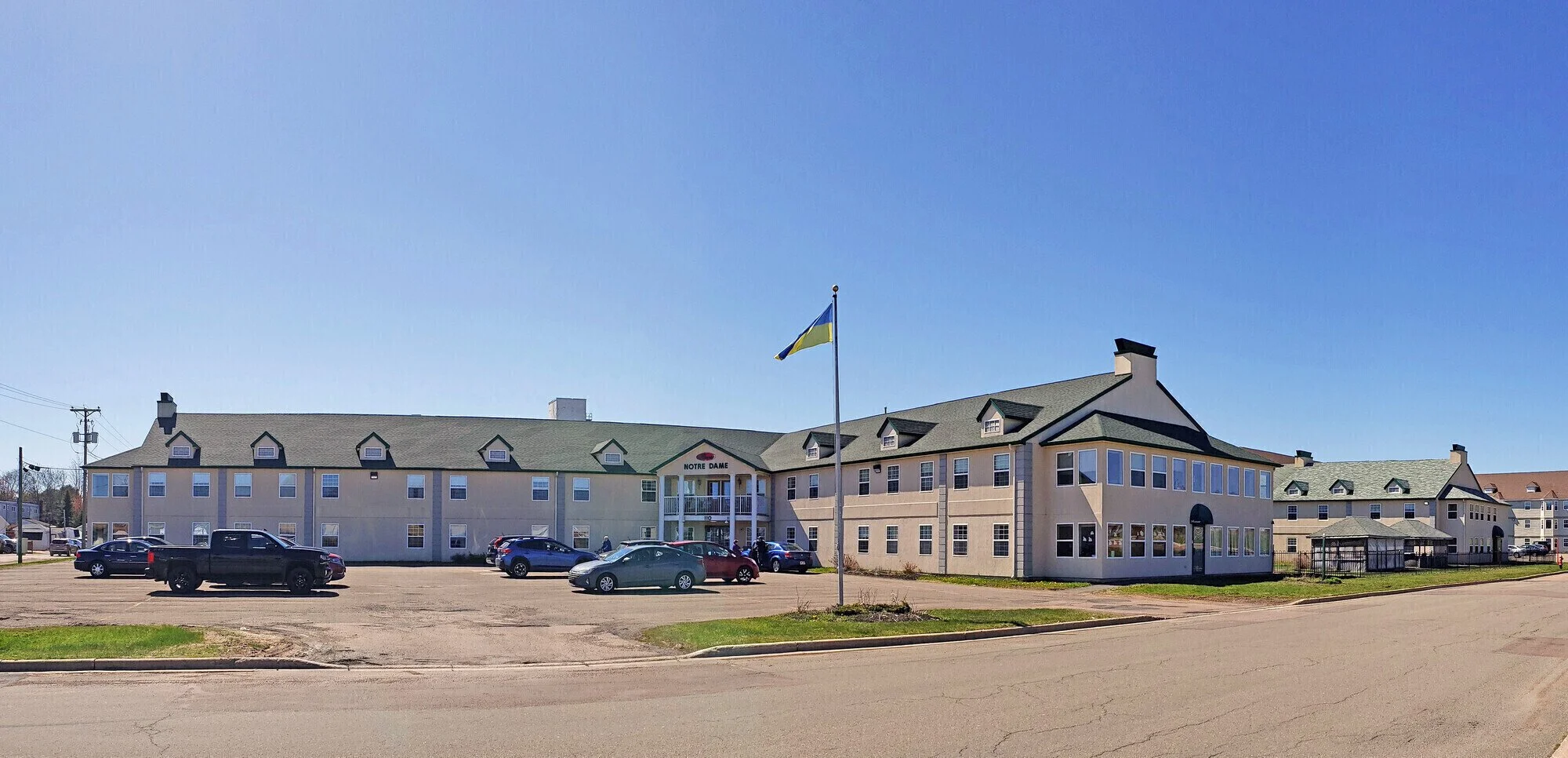 A large beige residential or hotel building with a green roof, multiple windows, and a sign that reads 'Notre Dame' at the entrance. In front, there is a parking lot with several parked cars and a flagpole with a flag waving. The sky is clear and blue.