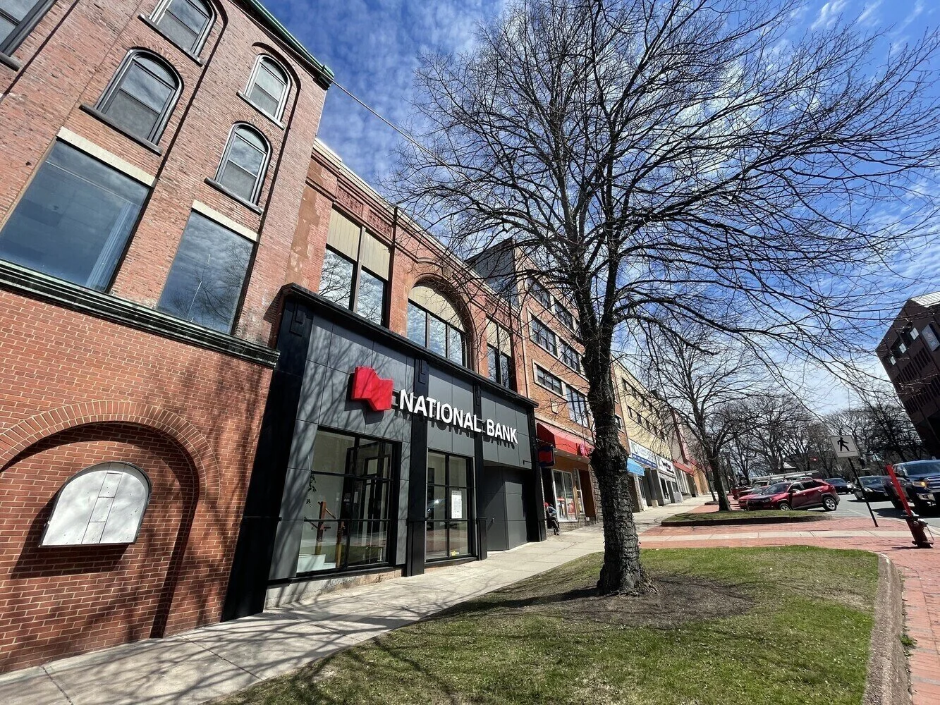 An urban street scene featuring a brick building with a National Bank branch, leafless trees, parked cars, and a clear blue sky.