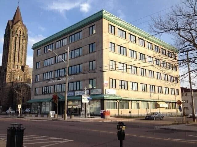 A multi-story office building with a beige exterior and green trim on the roof, located at a street corner with a parking lot and trees in front, and a church steeple visible in the background.