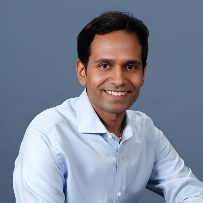 Portrait of a man in a light blue collared shirt smiling against a blue background.