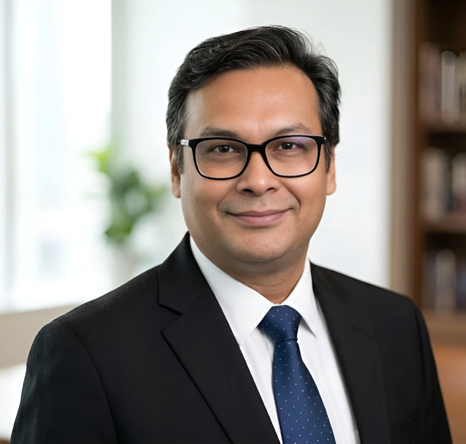 A man in a formal black suit, white shirt, and blue tie with white polka dots, wearing glasses, smiling, with bookshelves in the background.