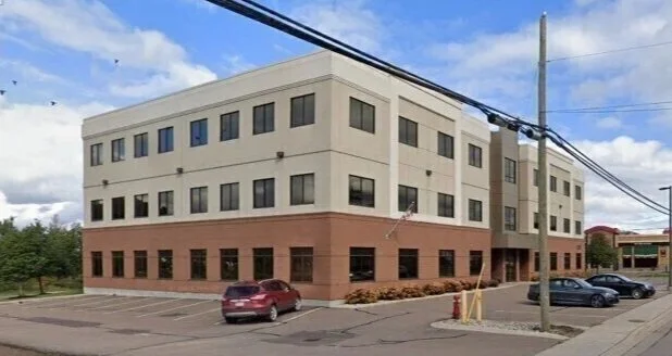 Four-story beige building with brick bottom floor, multiple windows, and a parking lot with a few parked cars in front.