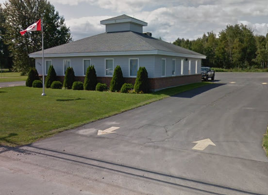 A light blue, single-story building with a gray gabled roof, surrounded by a manicured lawn and shrubs. A Canadian flag is flying on a flagpole near the building. An asphalt driveway with arrow markings leads to the building, with a parked vehicle visible on the right side. Trees and a partly cloudy sky are in the background.