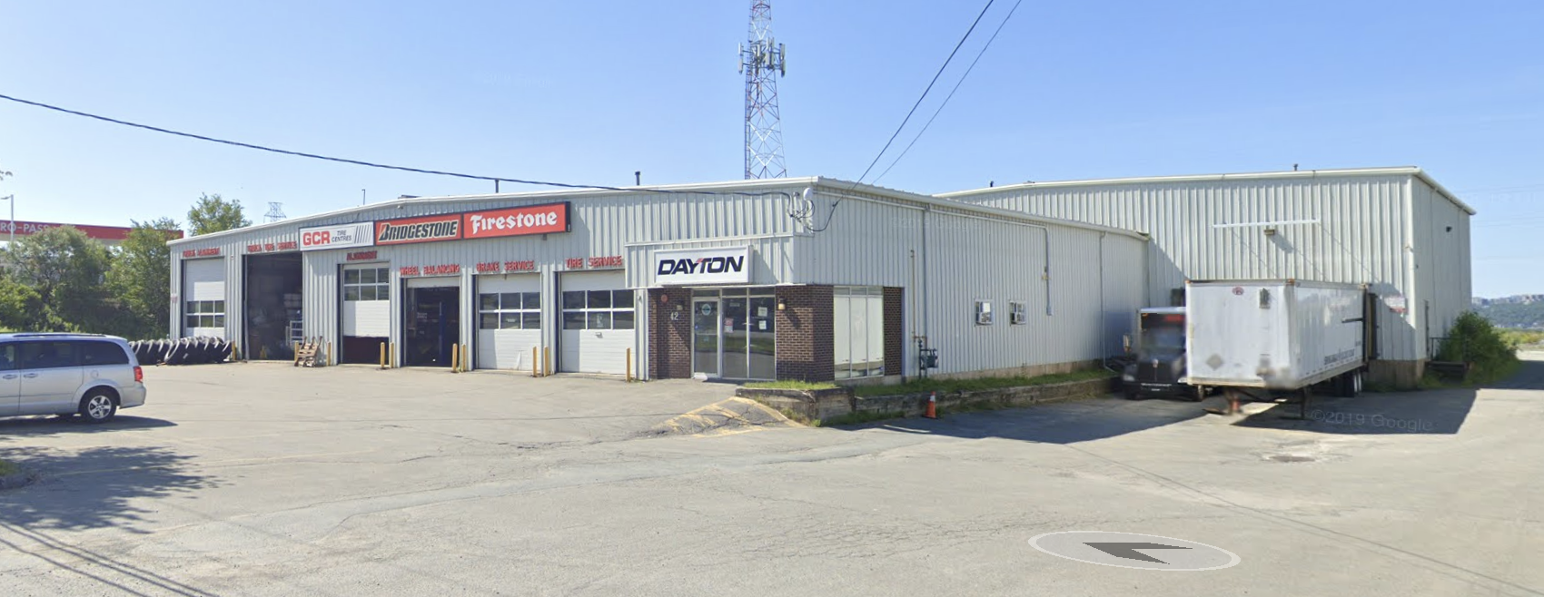 Automotive service garage with four bays, branded with Bridgestone and Firestone signs, and a small office entrance labeled Dayton, surrounded by a parking lot with a silver minivan and a delivery truck.