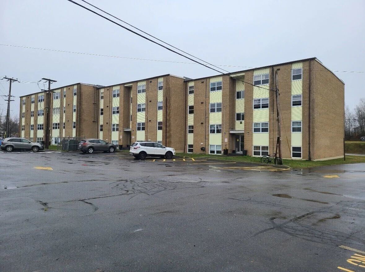 Multi-story residential apartment building with several parked cars and utility poles on a rainy day.