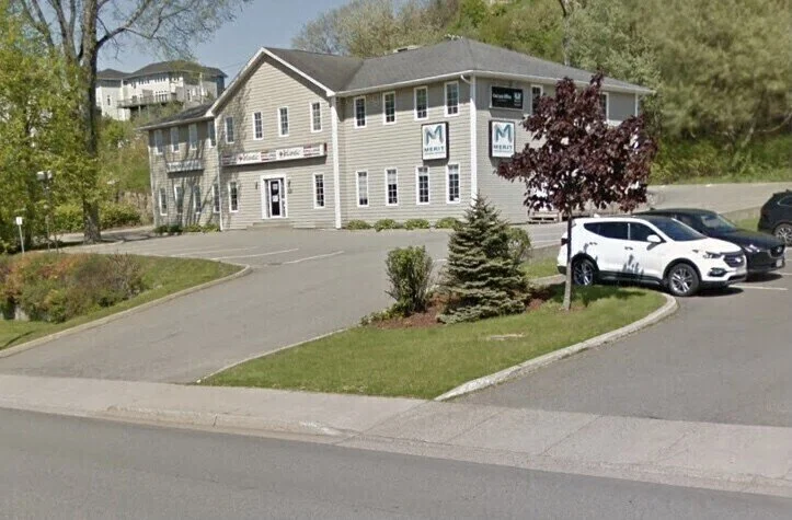 A two-story office building with gray siding, multiple windows, and signs that read 'Mercy.' There are landscaped areas with trees and bushes, and two white cars are parked in the lot in front.