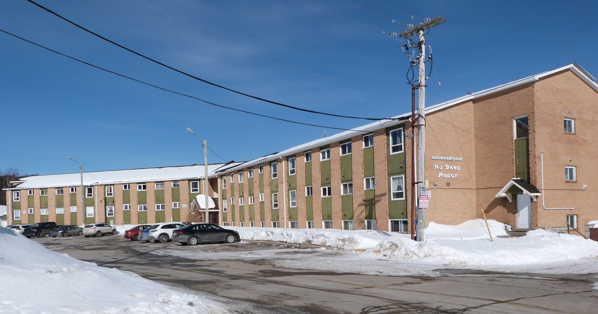 Snow-covered parking lot in front of a multi-story residential building with green and beige exterior, and a sign that reads "Nu Dawn Manor" under a clear blue sky.