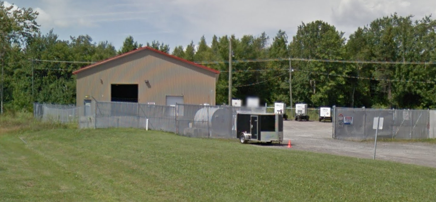 An industrial building with a fenced lot, several small trailers, and a grassy foreground.