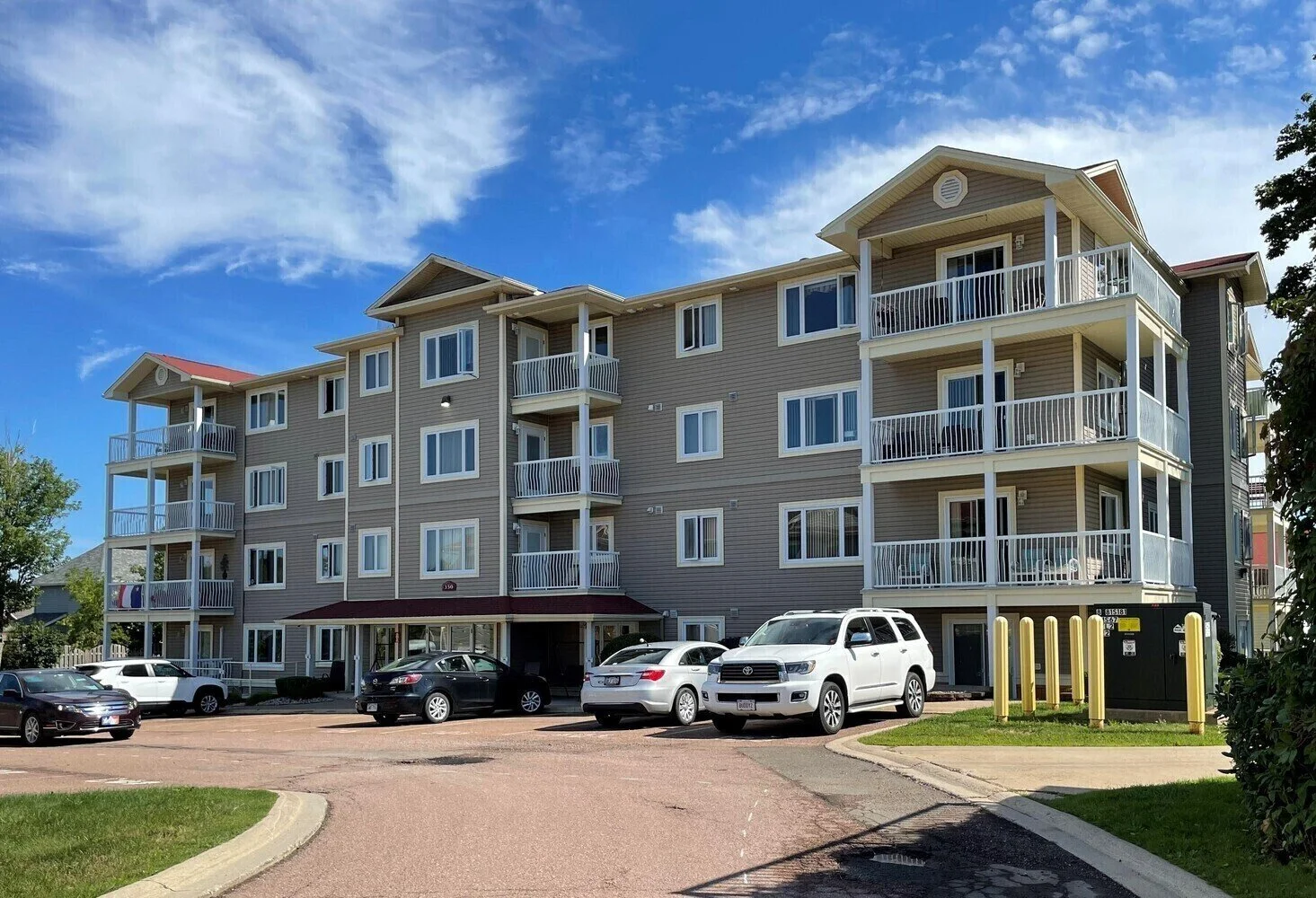 A multi-story apartment building with balconies and a parking lot in front, under a partly cloudy blue sky.