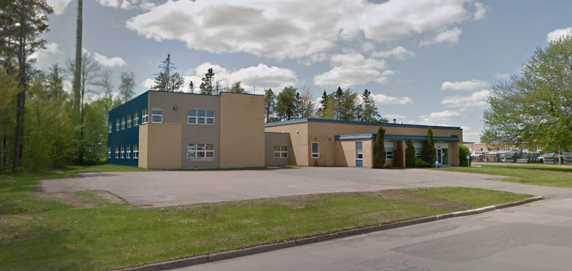 A beige and blue school building with a large parking lot and green grassy area in front.