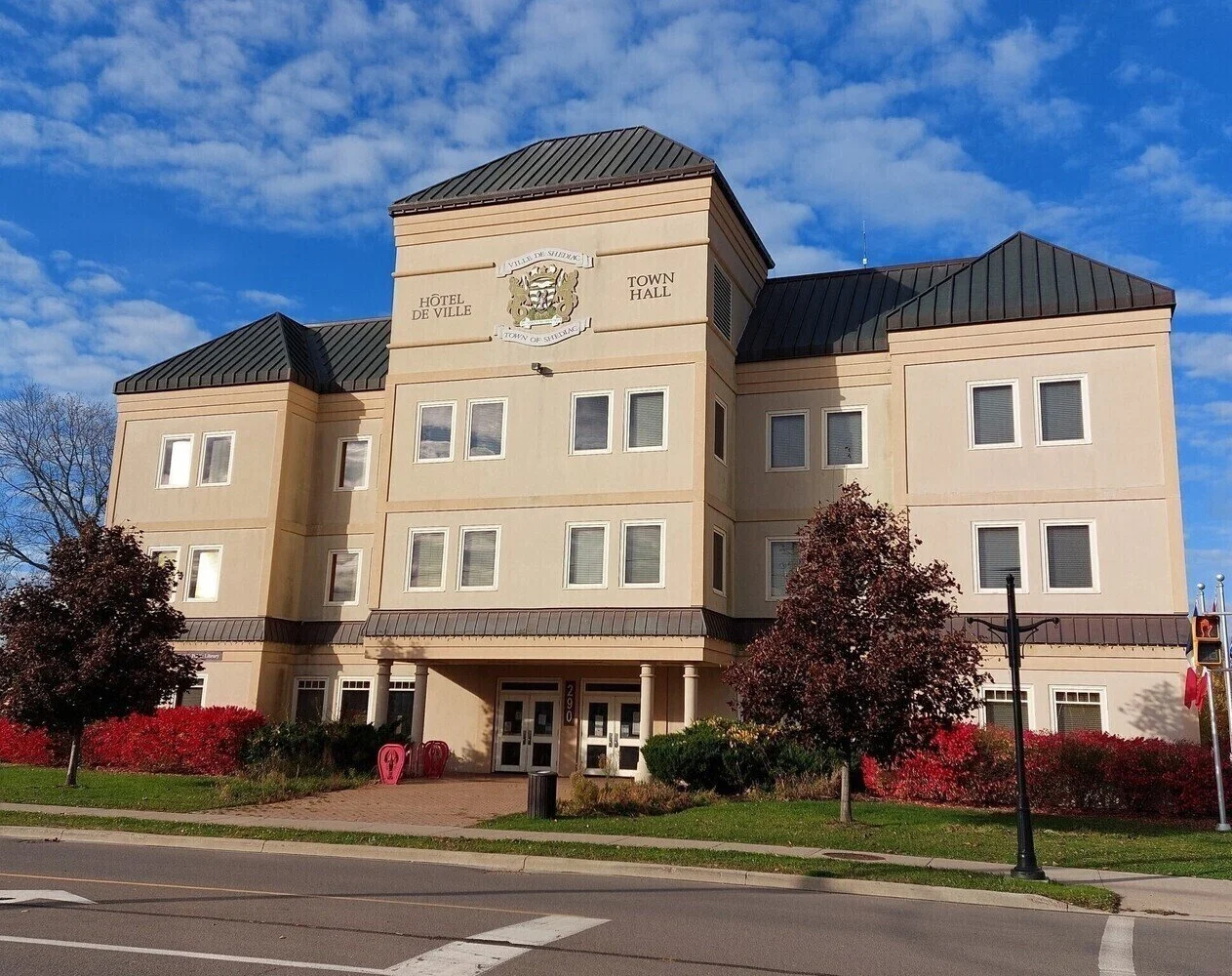 Three-story beige building labeled as Town Hall and Hôtel de Ville, with a crest sign on top, under a partly cloudy blue sky. There are red and purple bushes, trees, a sidewalk, and traffic signals in front.