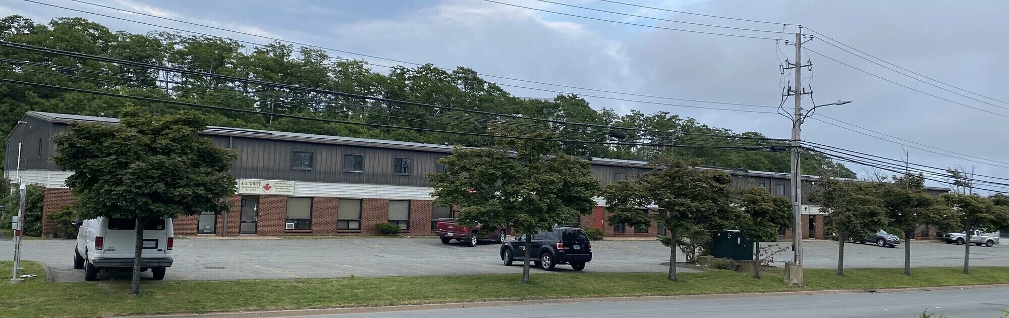 A parking lot in front of a mixed-use building with brick and dark siding, surrounded by trees, with power lines overhead and a cloudy sky.