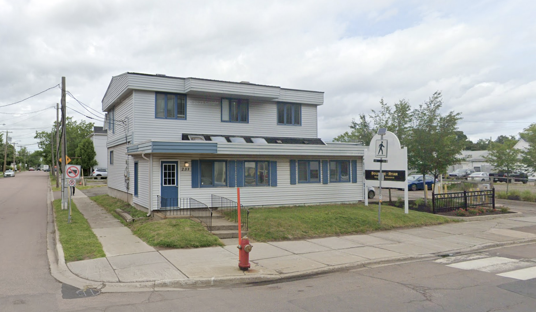 A three-story building with white siding, blue window shutters, and a small front porch, located on a sidewalk with a pedestrian crossing sign and a small garden with flowers in front. The street has parked cars and utility poles with power lines, under a cloudy sky.