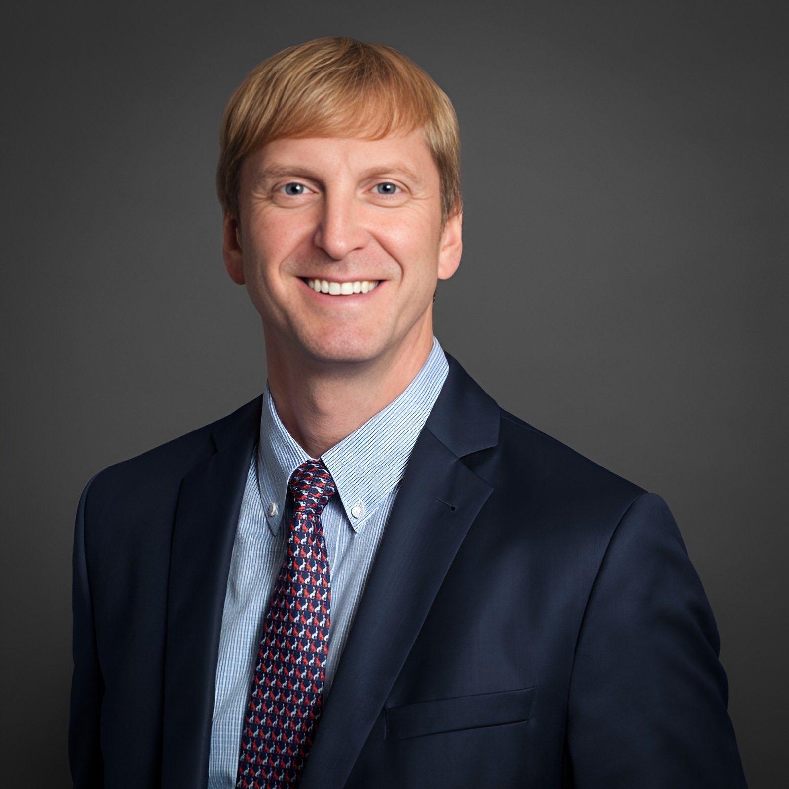 A man with blond hair wearing a dark suit, light blue dress shirt, and patterned tie, smiling against a plain gray background.