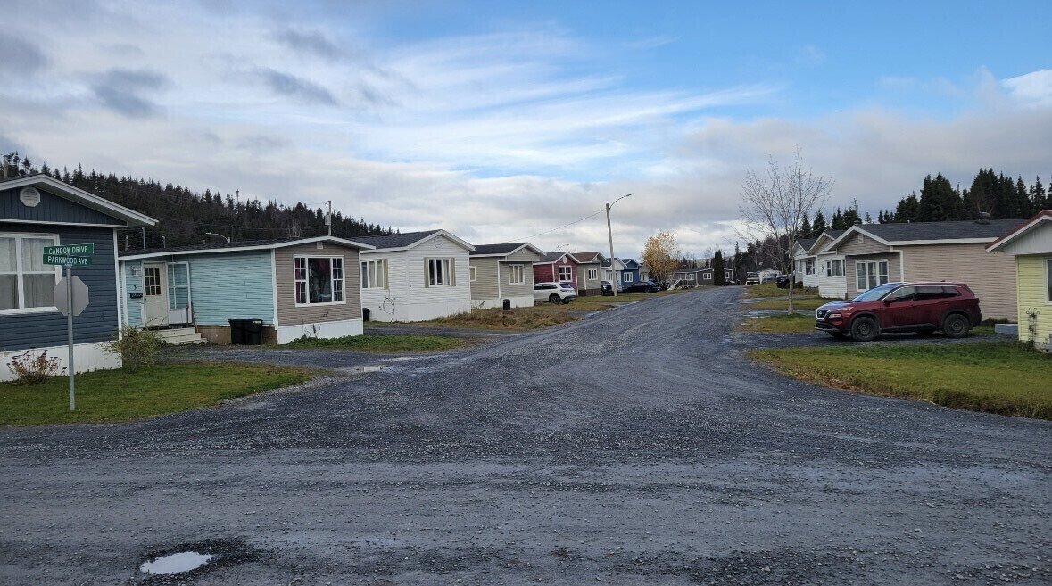 A residential neighborhood with small houses on a gravel street, some cars parked outside, and a cloudy sky above.