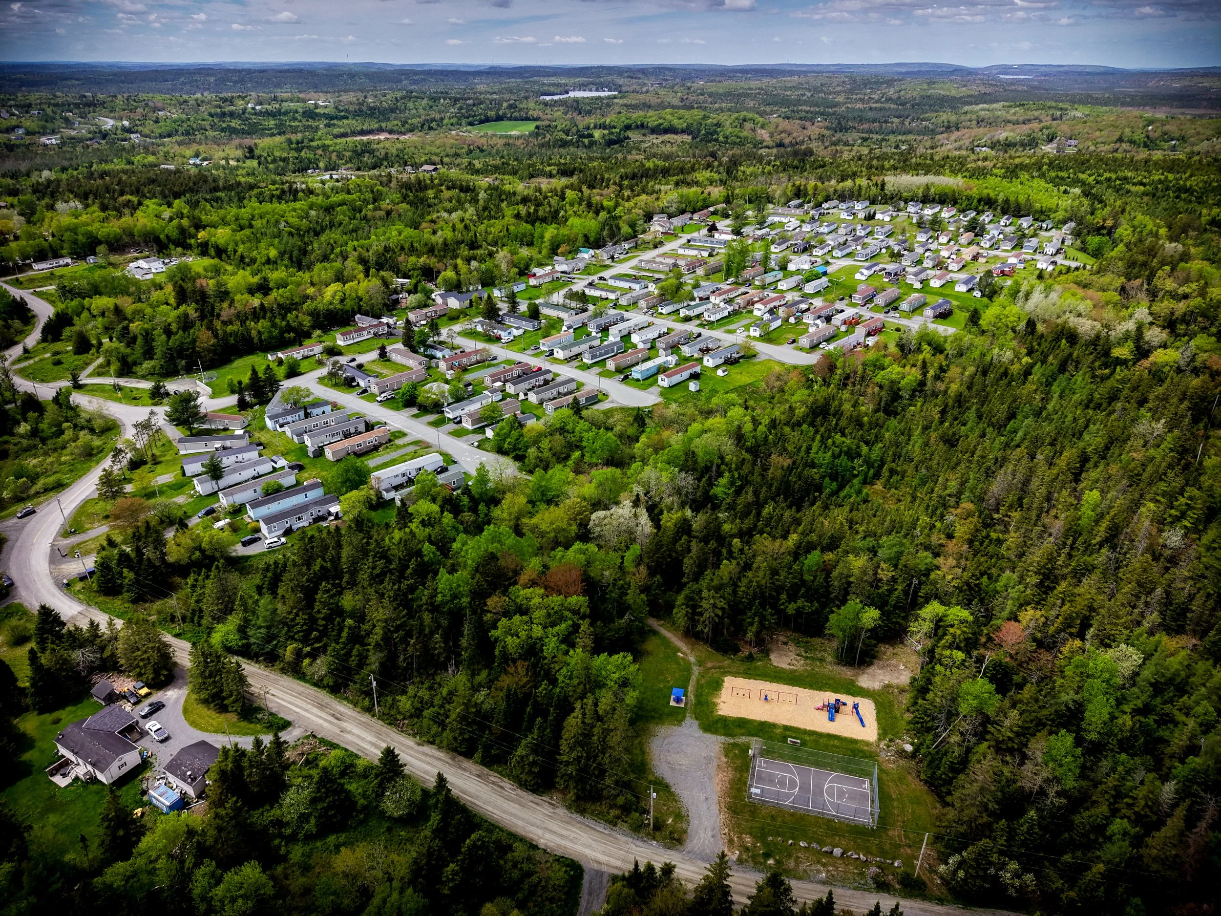Aerial view of a suburban neighborhood surrounded by dense green forests, with houses, roads, a playground, and a basketball court visible.