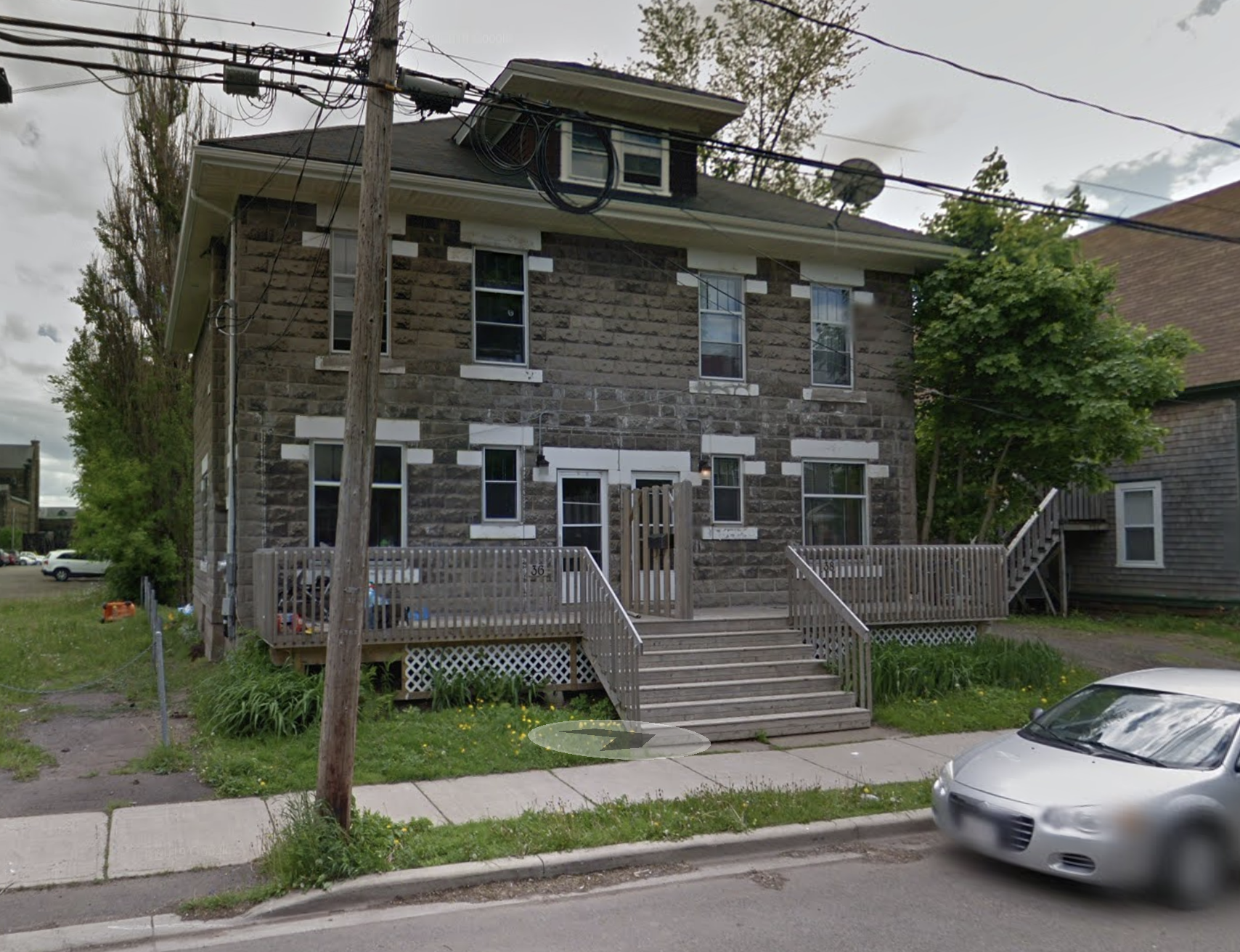 A three-story house with a stone exterior, multiple windows, a small front porch with stairs, a wooden fence, and a sidewalk. There is a utility pole with wires in front and a parked silver car on the street. Trees line the background.