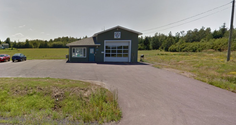 A small blue fire station with a garage door, surrounded by open green fields and parking lot, under a partly cloudy sky.