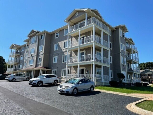 Multi-story apartment building with balconies, parked cars, and a clear blue sky.
